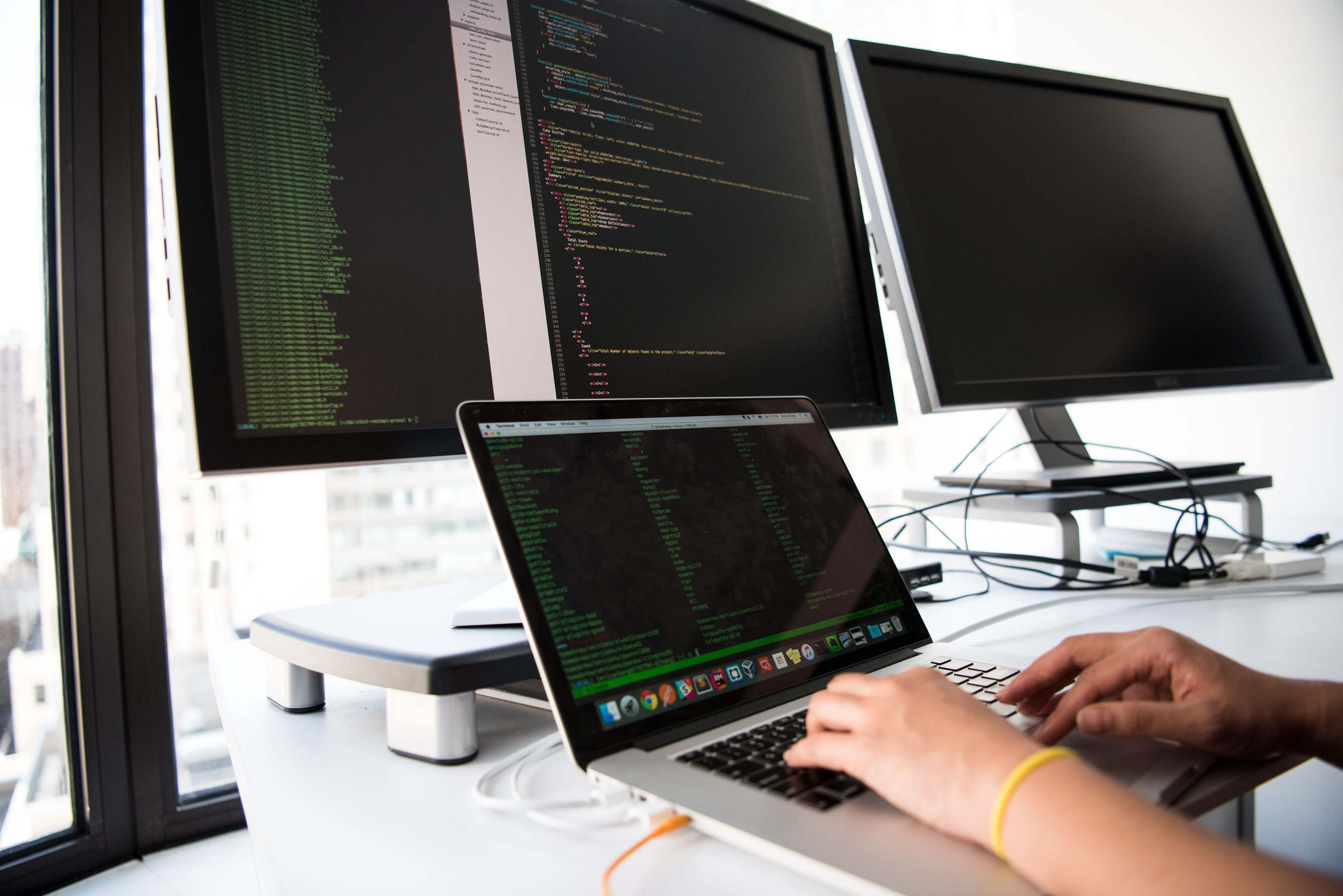 Hands typing on a laptop keyboard during software work, with code on a monitor in an office